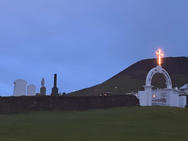 Picture of a cemetery at night. The entrance is a arch with a illuminated cross at the top.