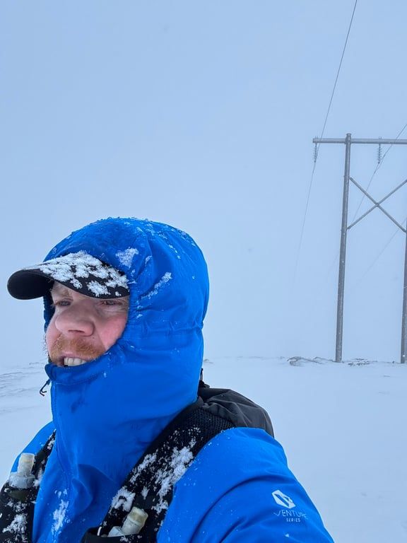 A selfie where I'm wearing a running west, a jacket zipped up to my mouth, hood up and a cap. The background is all white with snow and there. I'm also covered in snow.