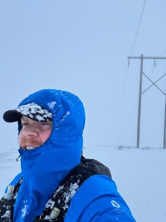 A selfie where I'm wearing a running west, a jacket zipped up to my mouth, hood up and a cap. The background is all white with snow and there. I'm also covered in snow.