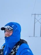 A selfie where I'm wearing a running west, a jacket zipped up to my mouth, hood up and a cap. The background is all white with snow and there. I'm also covered in snow.